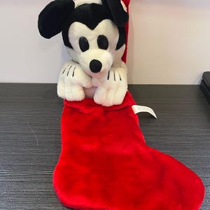 May include: A plush Mickey Mouse Christmas stocking. The stocking is red and the Mickey Mouse is white, black, and red. The plush toy is inside the stocking, with its head and arms visible. A red loop is attached to the top.