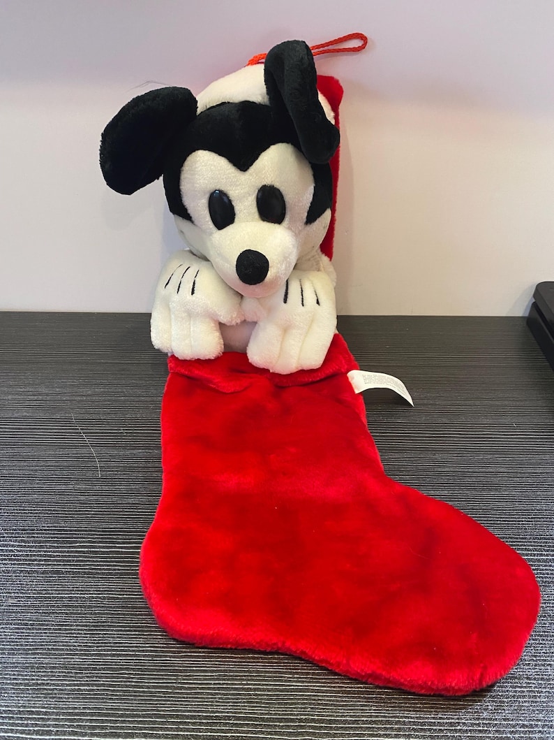 May include: A plush Mickey Mouse Christmas stocking. The stocking is red and the Mickey Mouse is white, black, and red. The plush toy is inside the stocking, with its head and arms visible. A red loop is attached to the top.