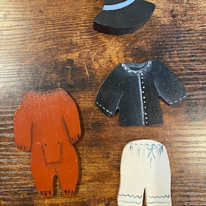 Puede incluir: Piezas de arte popular de madera que representan prendas de vestir. Incluye un sombrero negro con una banda azul, una chaqueta negra con costuras blancas, una figura en forma de oso marrón y pantalones cortos blanquecinos con detalles de olas azules.