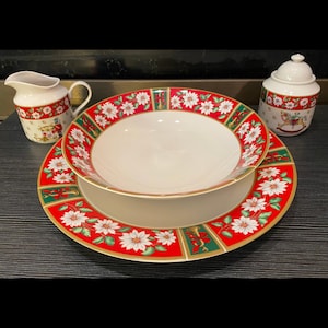 May include: A Christmas-themed porcelain dinnerware set featuring a red and green border with white poinsettias and gold trim. The set includes a large plate, a bowl, a creamer, and a sugar bowl with a lid. The sugar bowl has a rocking horse design.