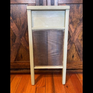 May include: A vintage-style washboard with a light green painted wooden frame. The washboard features a ribbed glass or plastic scrubbing surface. The design includes a rectangular top section and two legs.
