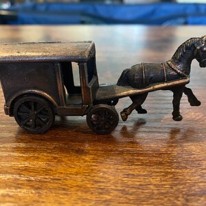 May include: A miniature antique-style horse-drawn carriage, crafted from dark metal. The carriage features a covered passenger area, spoked wheels, and a detailed horse figure. The overall design evokes a vintage aesthetic.
