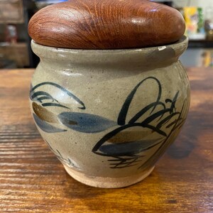 May include: A small, handcrafted ceramic jar with a wooden lid. The jar is beige with blue, brown, and black leaf designs. The lid is a smooth, polished brown. The jar is likely used for storage.