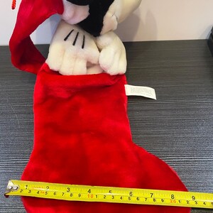 May include: A red plush Christmas stocking with a Mickey Mouse plush toy peeking out. The stocking measures approximately 10 inches long. The Mickey Mouse plush has black ears and a red Santa hat.