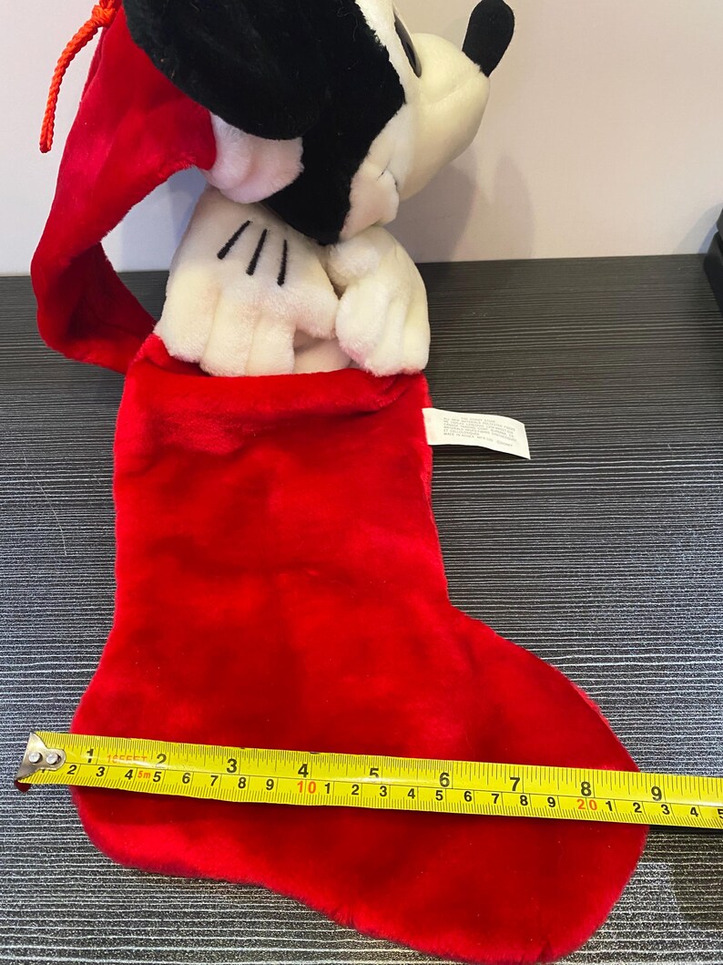 May include: A red plush Christmas stocking with a Mickey Mouse plush toy peeking out. The stocking measures approximately 10 inches long. The Mickey Mouse plush has black ears and a red Santa hat.