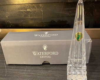 May include: A clear crystal obelisk-shaped decorative piece with a green and gold sticker. The obelisk is next to a gray box with the words "WATERFORD CRYSTAL" printed on it. The obelisk has a geometric design at the base.
