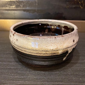 May include: A handmade ceramic bowl with a speckled, off-white rim and a dark brown interior. The bowl has a black base and a glossy finish, perfect for serving food or as a decorative piece. The bowl's diameter is approximately 6 inches.