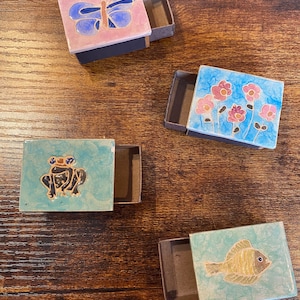 Könnte beinhalten: Vier dekorative Streichholzschachteln mit farbenfrohen Designs. Eine Schachtel zeigt einen Schmetterling, eine andere Blumen, eine dritte einen Frosch und die letzte einen Fisch. Die Schachteln sind geöffnet und zeigen das Streichholzfach.