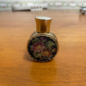 May include: A small, vintage perfume bottle with a gold-colored cap and a floral tapestry design. The bottle's body is round, with a black background and colorful flowers. The cap is a light gold color.