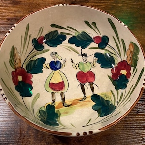 May include: A hand-painted ceramic bowl with a cream-colored interior and a brown rim. The bowl features a folk art design of two figures and floral elements in red, green, and blue. The bowl measures approximately 12 inches in diameter.