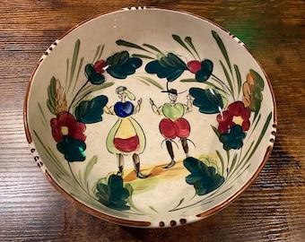 May include: A hand-painted ceramic bowl with a cream-colored interior and a brown rim. The bowl features a folk art design of two figures and floral elements in red, green, and blue. The bowl measures approximately 12 inches in diameter.