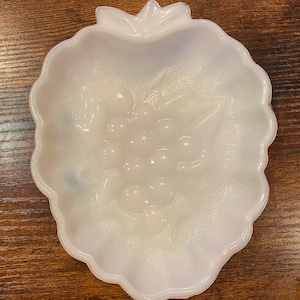 Puede incluir: Un plato de vidrio blanco opalino con forma de fresa. El plato presenta un diseño en relieve de uvas y hojas en el centro. Los bordes son festoneados y el plato está sobre una superficie de madera.