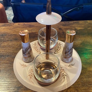 Puede incluir: Un juego de servir vintage con una bandeja rosa pálido, con dos saleros, dos vasos de vidrio transparente y un soporte central. El juego tiene detalles dorados y un patrón de trigo.
