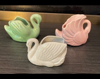 May include: Three ceramic swan ornaments in pastel shades: light green, pink, and white. Each swan has a hollowed-out area, suggesting they could be used as small planters or decorative containers. The swans are arranged on a dark surface.