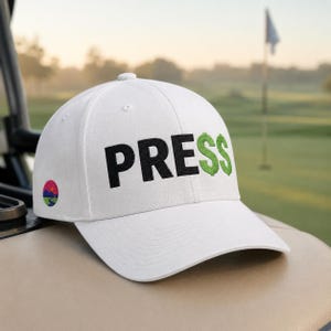 Könnte beinhalten: Weiße Baseballkappe mit dem Wort "PRESS" in Schwarz und Grün gestickt. Das "S" ist ein Dollarzeichen. Auf der Seite befindet sich ein farbenfrohes kreisförmiges Logo. Die Kappe liegt auf einem Golfwagen.