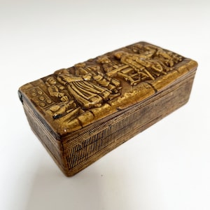 May include: A rectangular wooden box with a raised, detailed scene of people in a village setting. The box is a dark brown color and has a textured surface.
