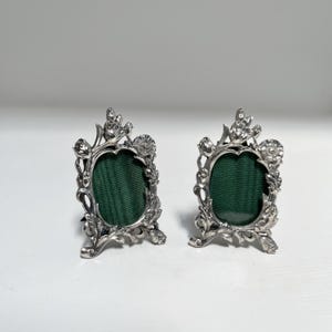 Può includere: Due piccole cornici portafoto in metallo color argento di forma ovale, caratterizzate da dettagli in rilievo a forma di fiori e foglie lungo i bordi. Ogni cornice presenta un inserto in tessuto verde scuro a coste orizzontali.