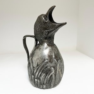 May include: A dark metallic pitcher shaped like a bird with its beak open. The body of the pitcher features a textured relief pattern of tall grass or reeds. It includes a curved handle attached to the side, with a weathered, aged finish.