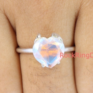 Pu&ograve; includere: Un anello d'argento con una pietra preziosa iridiscente a forma di cuore. L'anello &egrave; su un dito.