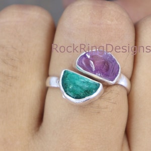 May include: A silver ring featuring two half-circle gemstones. The left side is a green emerald and the right side is a purple amethyst. The ring is worn on a finger. The text "RockRingDesigns" is visible on the ring.