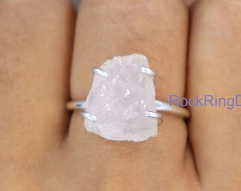 May include: A silver ring with a raw, pink rose quartz stone. The stone is set in a simple, wire-wrapped design. The ring is shown on a person's finger.