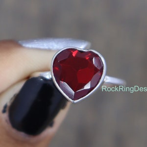 May include: A silver ring with a red heart-shaped gemstone. The ring is set in a simple band and the gemstone is faceted.