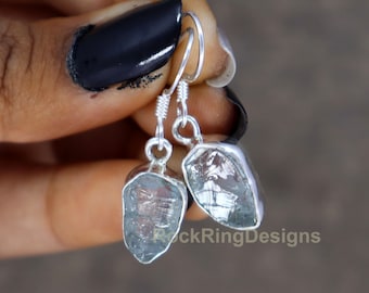 Può includere: Orecchini d'argento con pietre di acquamarina grezze. Le pietre sono trasparenti e hanno una tonalità blu chiaro. Gli orecchini sono appesi a un gancio d'argento.