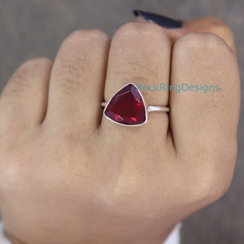 Puede incluir: Un anillo de plata con una piedra roja triangular. El anillo est&aacute; en el dedo de una persona.