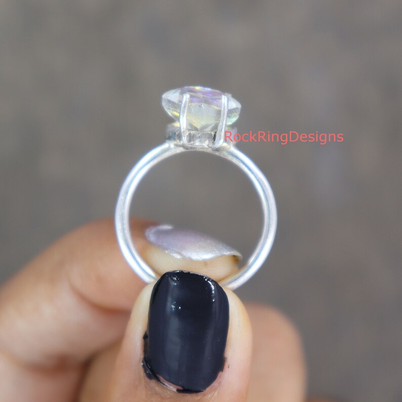 Pu&ograve; includere: Un anello d'argento con una pietra preziosa trasparente e iridescente. L'anello &egrave; incastonato su una fascia semplice ed &egrave; tenuto da una mano con smalto nero.