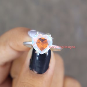 Pu&ograve; includere: Un anello d'argento con una pietra preziosa di opale iridescente a forma di cuore. L'opale presenta un arcobaleno di colori, tra cui arancione, rosso e blu.
