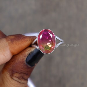May include: A silver ring with a pink and green oval gemstone. The ring has a split shank design. RockRingDesigns is printed on the band.