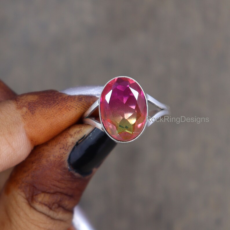 May include: A silver ring with a pink and green oval gemstone. The ring has a split shank design. RockRingDesigns is printed on the band.