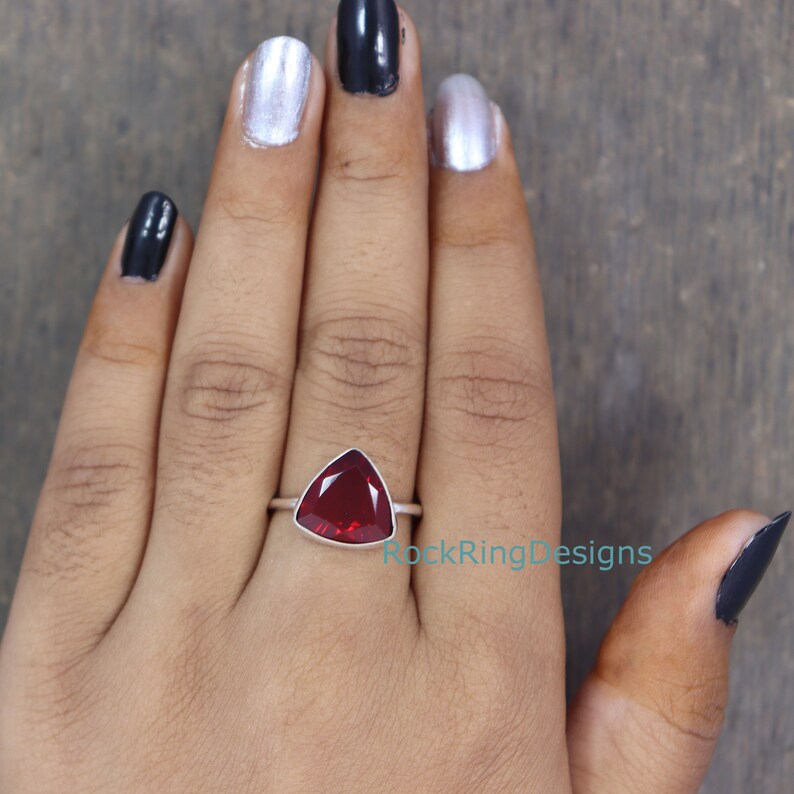 Puede incluir: Un anillo de plata con una piedra roja triangular. El anillo est&aacute; en el dedo de una persona.