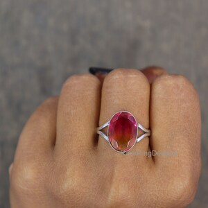 May include: A silver ring with a pink and green oval gemstone. The ring is on a person's finger.