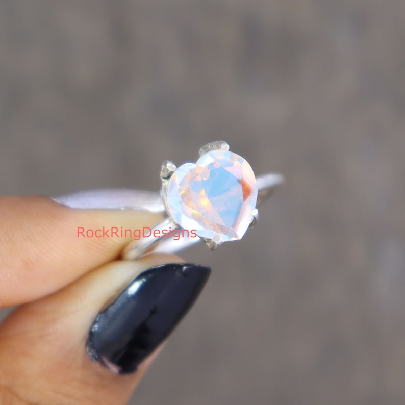 Pu&ograve; includere: Un anello d'argento con una pietra preziosa di opale trasparente a forma di cuore. L'anello &egrave; indossato su un dito con smalto nero.