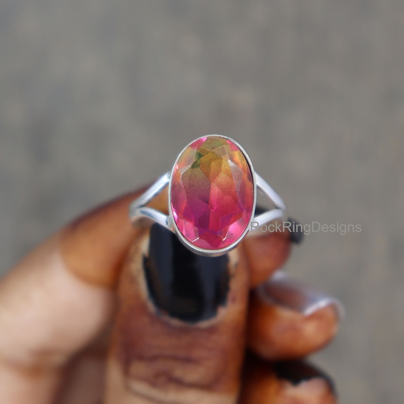 May include: A silver ring with a large oval-shaped gemstone. The gemstone is a deep pink color with a gradient of yellow and orange hues. The ring is set in a simple band with a split design.