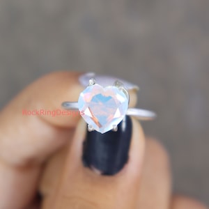 Pu&ograve; includere: Un anello d'argento con una pietra preziosa di opale iridescente a forma di cuore. L'anello &egrave; incastonato su una fascia semplice.