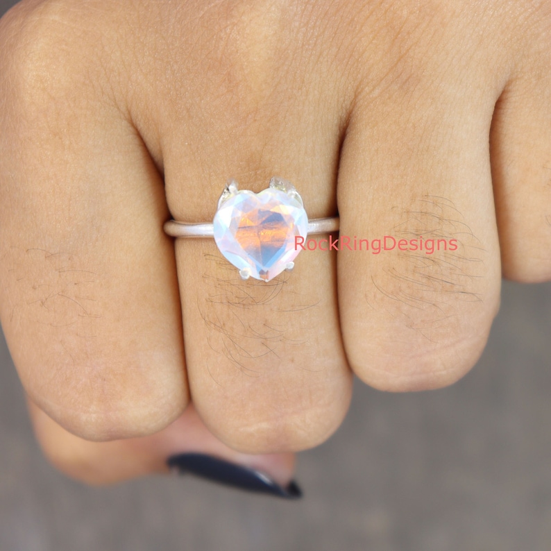 Pu&ograve; includere: Un anello d'argento con una pietra preziosa iridiscente a forma di cuore. L'anello &egrave; su un dito.