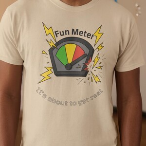 May include: Beige t-shirt with a graphic of a fun meter, featuring a gauge with green, yellow, and red sections. The text "Fun Meter" and "It's about to get real" are also printed on the shirt.