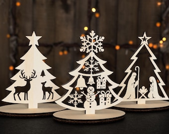 May include: Three wooden Christmas tree ornaments with different designs. The first tree has a deer and a smaller deer on the front. The second tree has a snowflake, a snowman, and a bell on the front. The third tree has a star on top and a nativity scene on the front.