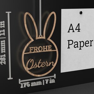 Może przedstawiać: Drewniana ozdoba wielkanocna w kształcie kr&oacute;lika z napisem "Frohe Ostern" w k&oacute;łku. Ozdoba ma 176 mm szerokości i 281 mm wysokości.
