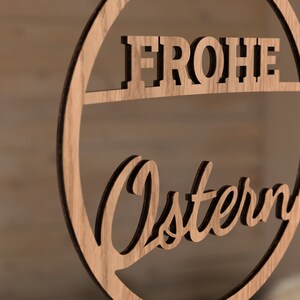 Może przedstawiać: Drewniane koło z wyciętymi słowami "Frohe Ostern" w ozdobnym kroju pisma. Koło jest jasnobrązowe i ma naturalne usłojenie drewna.