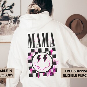 May include: White hoodie with a black and white checkered design and a pink smiley face with lightning bolts. The word "MAMA" is printed in black above the design.