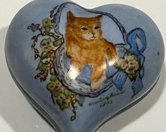 May include: A blue heart-shaped ceramic trinket box with a painted image of an orange tabby cat in a blue ribboned basket with flowers. The box is signed "Marisabel 1983".