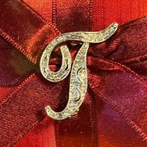 Pode incluir: Um broche de prata em forma de letra T com strass sobre um fundo de tecido vermelho e bordô.