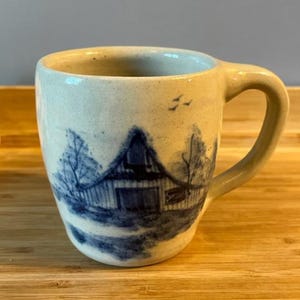 以下が含まれることがあります： クリーム色の陶器マグカップ。青い納屋の風景が描かれています。取っ手はカーブしており、素朴で手作りの外観です。納屋の風景には木や鳥が含まれています。