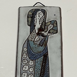 May include: A ceramic wall hanging depicting a stylized image of the Virgin Mary holding a baby Jesus. The artwork is in shades of blue, gray, and white with black accents.