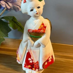 Puede incluir: Muñeca geisha de cerámica con kimono blanco y estampados florales rojos. La muñeca tiene un peinado detallado con detalles rojos y dorados, y un adorno verde. Está sobre una superficie de madera.