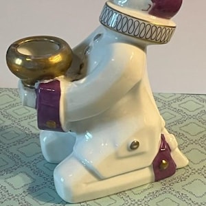 May include: A white ceramic figurine of a kneeling person with a gold-trimmed collar and a purple head. The figurine is holding a gold-colored bowl. The figure has purple accents on the sleeves and knees.