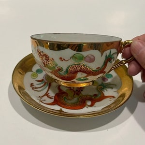 May include: A white teacup and saucer with a gold rim and a red and green dragon design. The cup has a gold handle.
